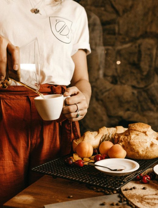 A woman pours coffee into a cup, surrounded by fresh bread and fruits in a cozy café setting.
